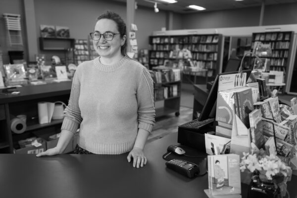Black-and-white photograph of a woman standing at a counter in a bookstore. The bookshelves are visible behind her. She wears glasses and smiles at the photographer.