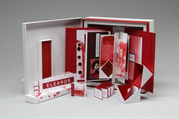 Photograph shows a three-dimensional artwork. The artwork shows an open book without pages. There is a red label reading "Eleanor" on the lower left side of the book. There are red and white pages bent and folded in patterns.