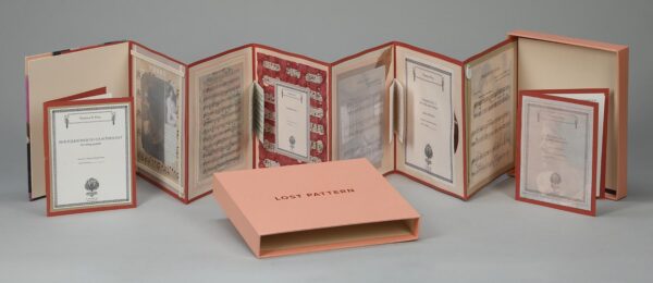 Photograph shows an unfolding book. There is text written on the pages. A book with a pink cover reads "Lost Pattern"