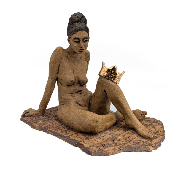 Ceramic sculpture of a nude woman, seated and leaning back on her extended arms. A gold crown is perched on one knee. She looks down with a downcast expression.