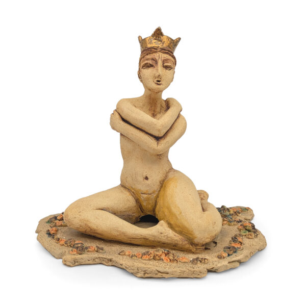 Ceramic sculpture of a nude woman seated and wrapping her arms around herself. She wears a gold crown. There are small ceramic plants on the ground around her.