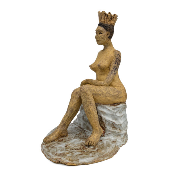 Ceramic sculpture of a nude woman, seated and turned to the side. She wears a gold crown. There is black patterned script down her arm.
