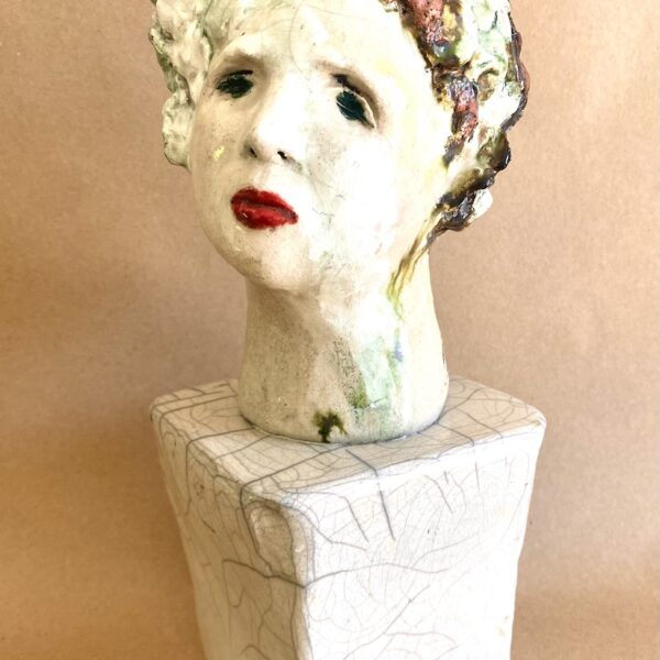 Bust on a rough rectangular base. The pale white head has a female face with red lips and reddish-brown hair. There are light green leaves adorning the hair.