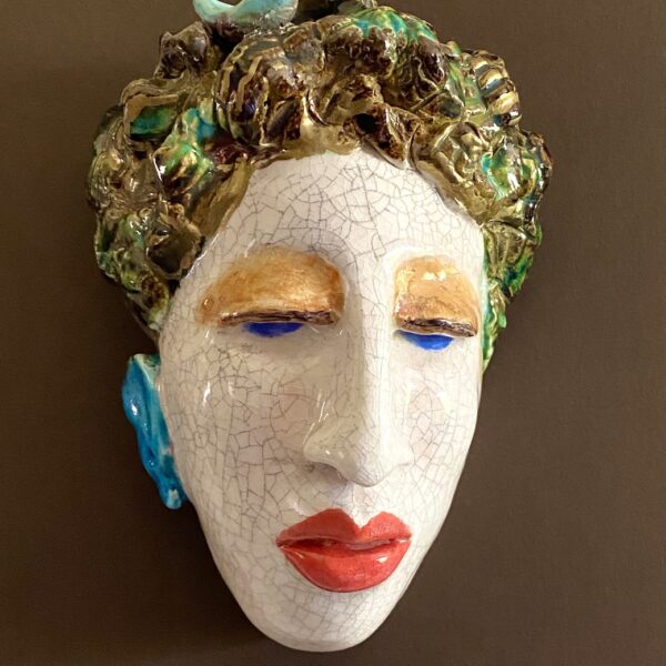 Ceramic sculpture of a head with red lips and brown hair. The eyes are blue with vivid brownish eyeshadow. The face has a blue ear visible. The hair is adorned with greenish accents and the small blue figure of a bird.