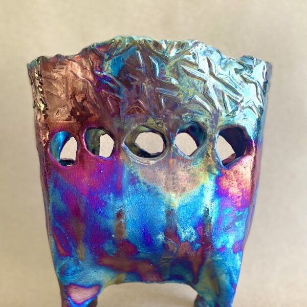 Ceramic vessel on two feet. There are holes in a line through the center of the piece. It is a bright iridescent rainbow color.