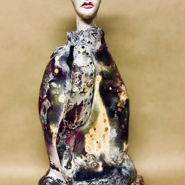 Ceramic figure wearing elaborate robes. The figure has a pale white face and red lips. She wears a crown with wire.