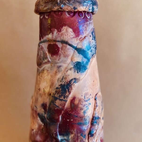 Cylindrical ceramic sculpture on a base with two feet. The sculpture has abstract colors of red, blue, white and pale pink. A lid and handle sit atop the sculpture.