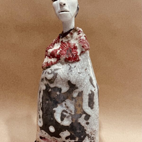 Elongated ceramic figure with a pale white head. The figure is wrapped in a garment with a large reddish collar. She wears a headdress with red accents.