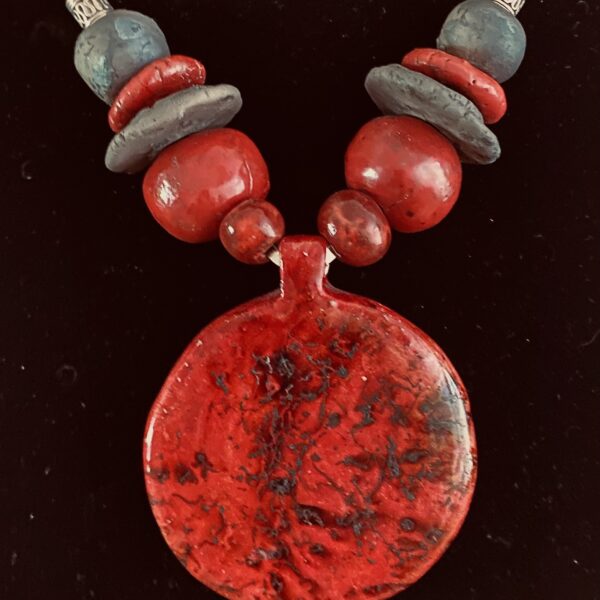 Necklace of large irregular flat and round ceramic beads, with a large flat circular pendant at its center. The beads are gray and vivid red hues.