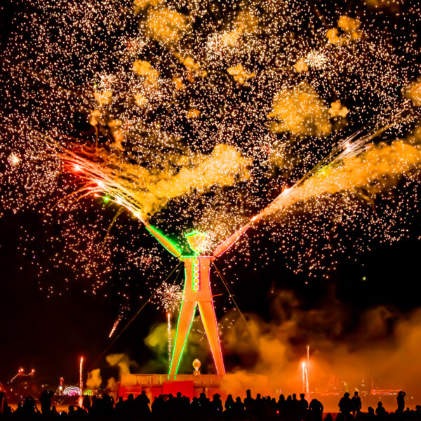 Photograph shows a large featureless statue of a man lit up against a night sky. Sparks and flames leap around it.