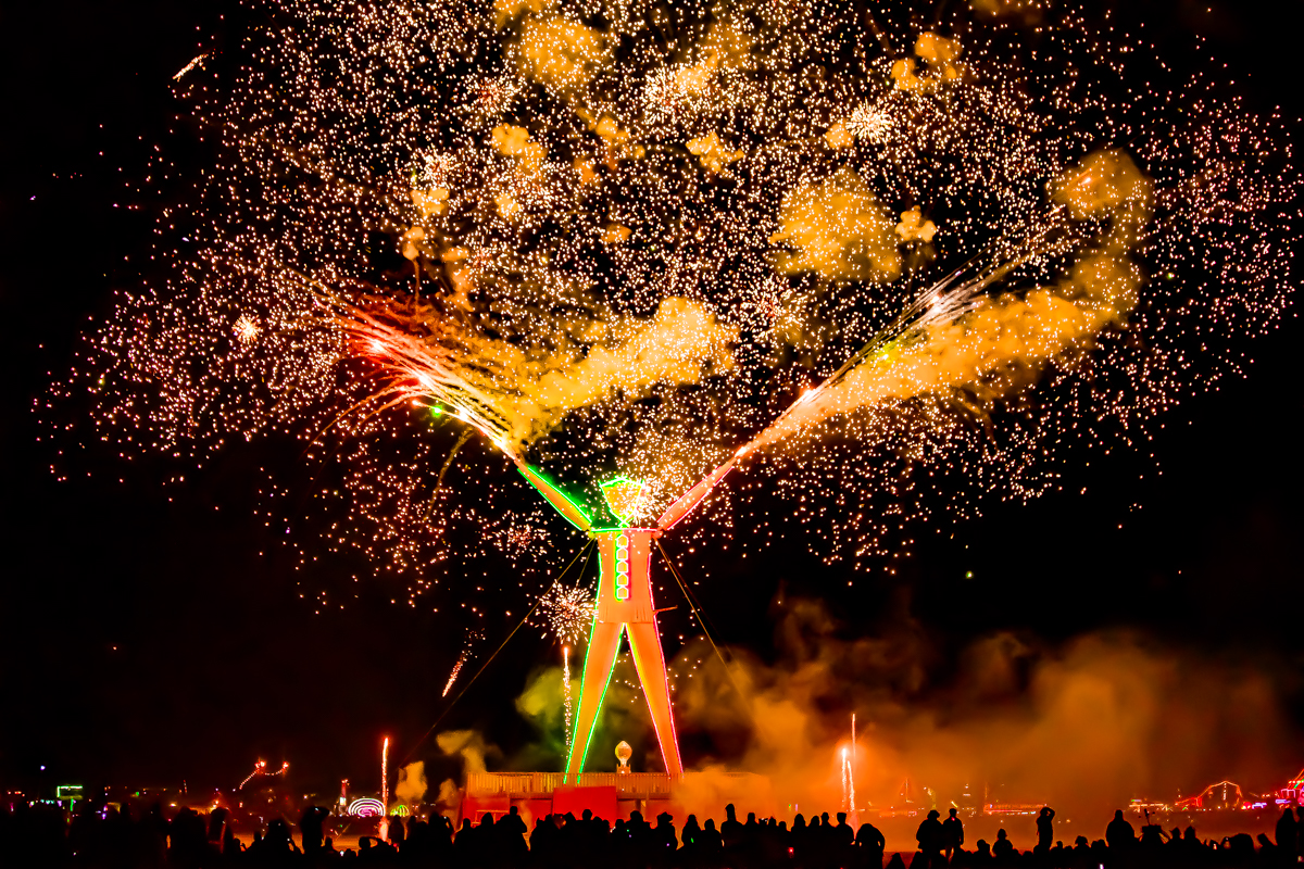 Photograph shows a large featureless statue of a man lit up against a night sky. Sparks and flames leap around it.
