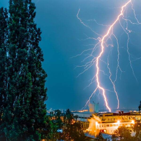 Photograph shows lightning striking a building in the distance. A tree is in the foreground. The sky is stormy.