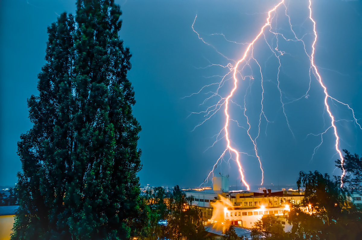Photograph shows lightning striking a building in the distance. A tree is in the foreground. The sky is stormy.