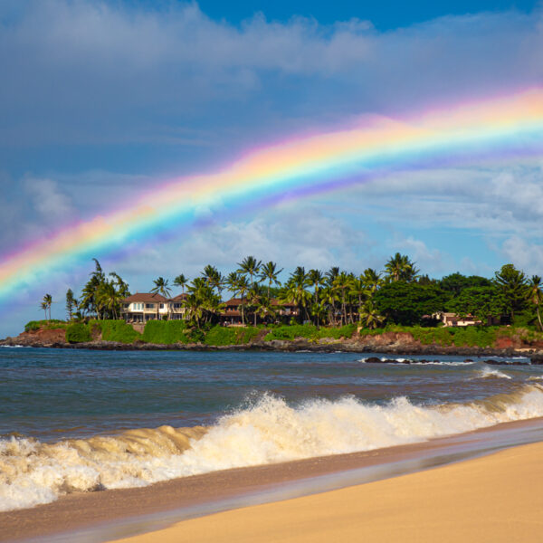 Photograph showing a rainbow over a beach with a wave crashing on the sand.