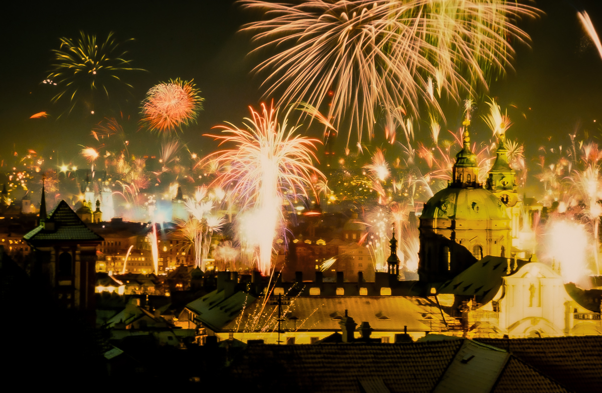 Photograph shows fireworks exploding against a dark night sky over buildings and rivers. The fireworks are shades of gold, illuminating the city below.