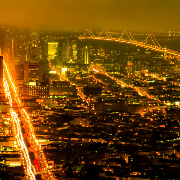 Photograph shows traffic on a freeway from the air at night. Red and white lights stream across the freeway. A city is lit up below. A bridge is visible crossing the water in the distance.