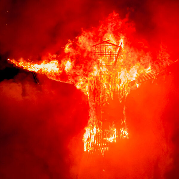 Photograph of a large statue of a man. The statue is on fire with brilliant orange-red flames against a dark night sky.