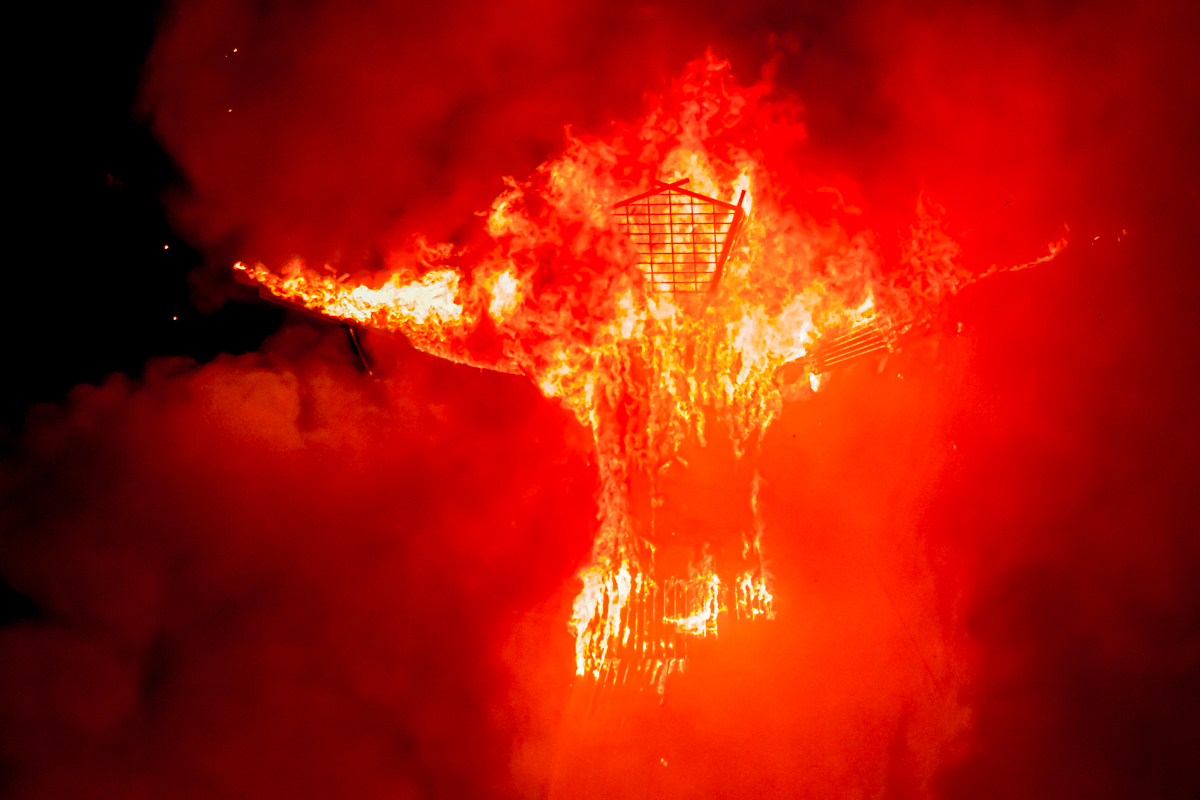 Photograph of a large statue of a man. The statue is on fire with brilliant orange-red flames against a dark night sky.