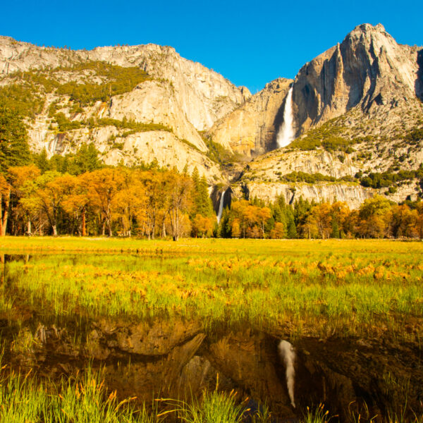 Photograph showing a distant waterfall down a rock face. The image reflects in the river below. The sky is a brilliant blue.