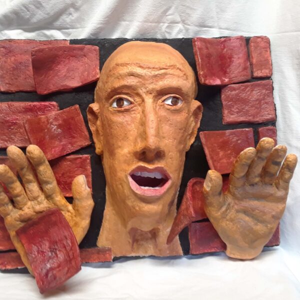 Photography showing a 3D art piece. A bald-headed man peers from a broken red brick wall. His hands push bricks away.