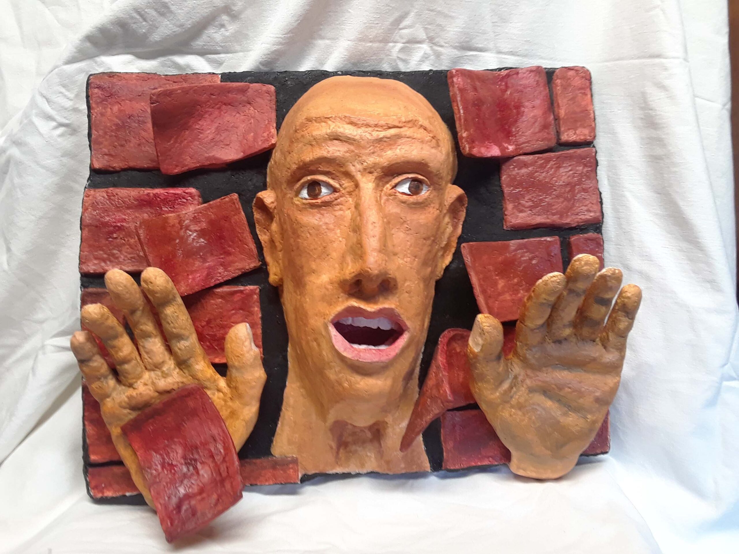 Photography showing a 3D art piece. A bald-headed man peers from a broken red brick wall. His hands push bricks away.