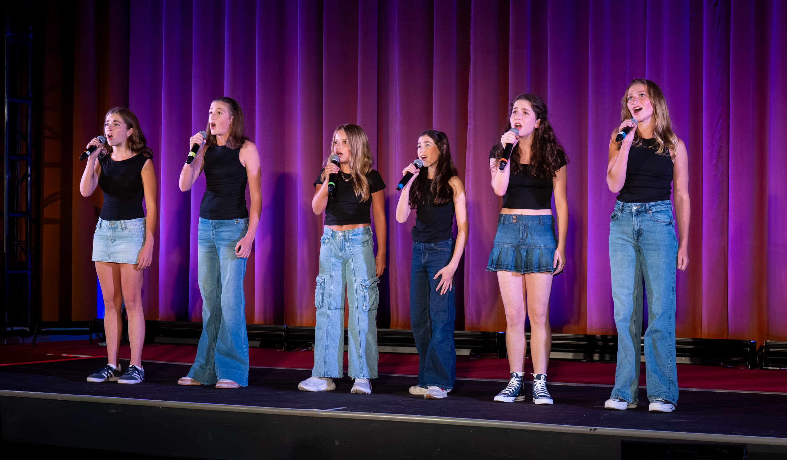 Photograph shows a group of girls singing into microphones on a stage