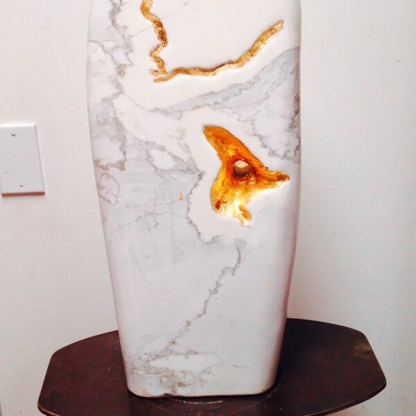 Photograph showing a white stone sculpture sitting on a wooden stand. The sculpture has abstract gold decorations.