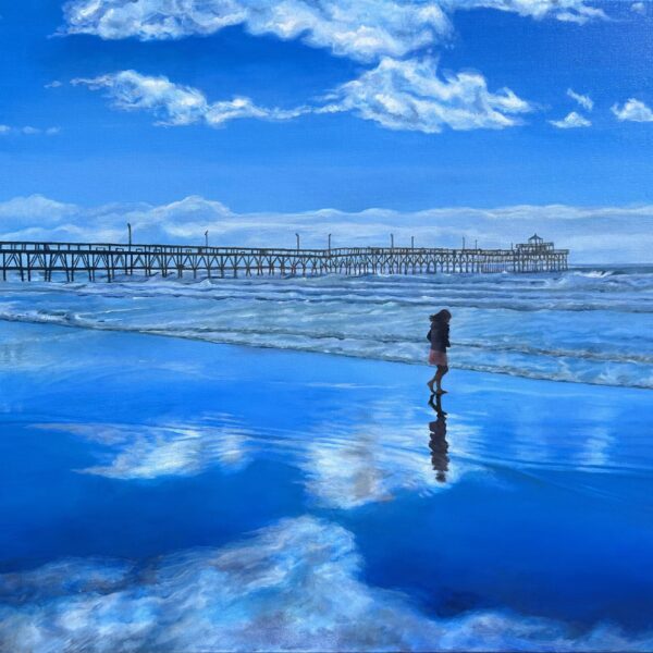 Image shows a figure walking along a beach. The water reflects the blue sky above. A pier is visible in the distance